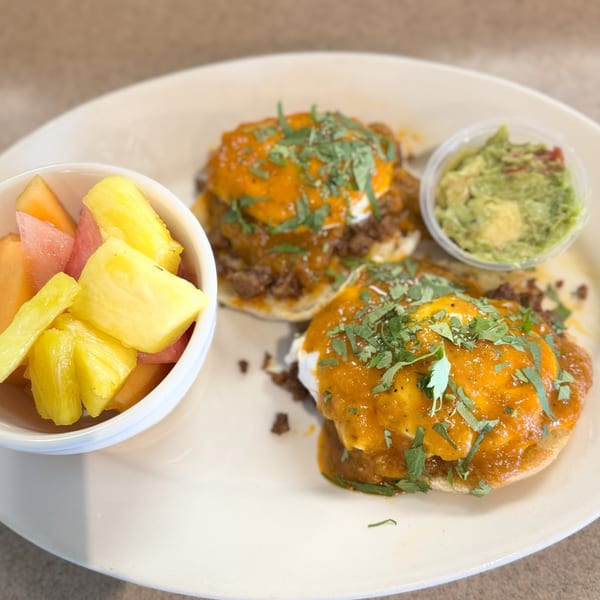 Two huevos rancheros with a side of fruit salad and guacamole on a white plate.