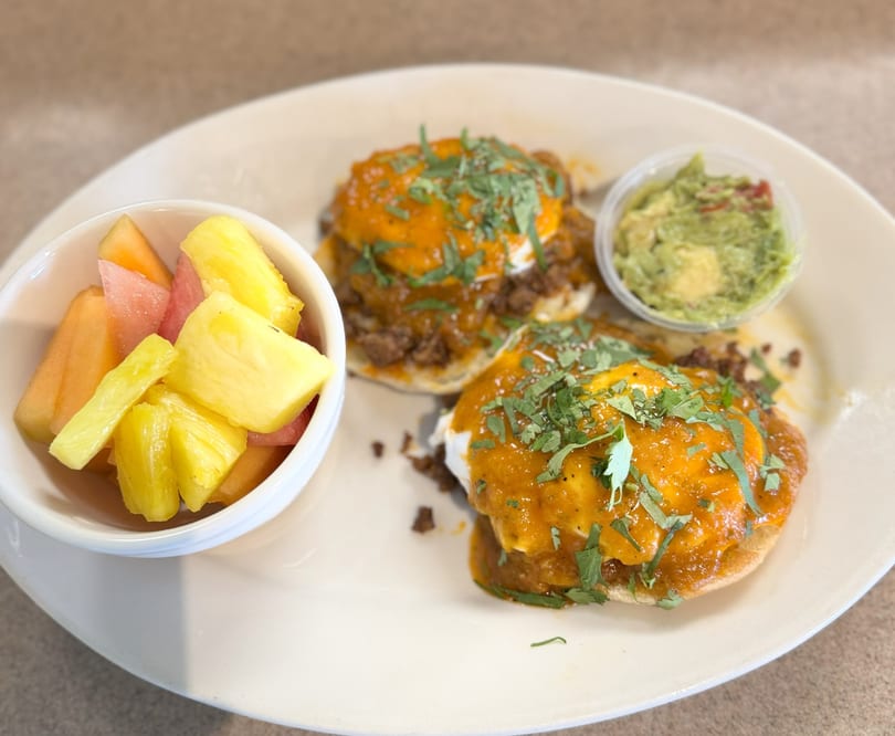 Two huevos rancheros with a side of fruit salad and guacamole on a white plate.